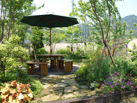 パラソル付きのテーブルとベンチを設けた、雑木と草花に囲まれたテラスガーデンの施工後全景。