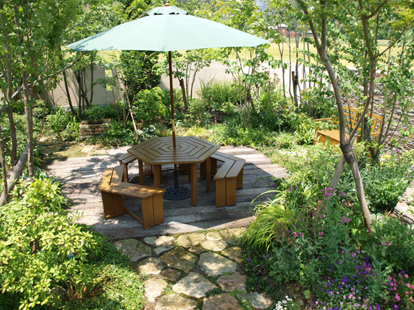 パラソルの下でくつろげるテーブルとベンチ、周囲を彩る草花と雑木。